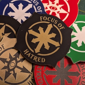 Op de afbeelding: Een verzameling cirkelvormige en schildvormige tokens met een sterrenpatroon en de woorden "Focus of Hatred". De tokens zijn in verschillende kleuren, waaronder zwart, goud, rood, blauw en groen. Het ontwerp bevat pijlen die naar buiten wijzen.