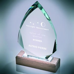 May include: A clear glass award with a faceted, teardrop-shaped top and a wooden base. The text "Twilight Recruitment," "Sales of the Quarter," "WINNER," and "ASTRID PIERRE" are etched into the glass. The award is approximately 8 inches tall.