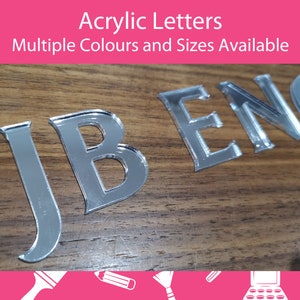 以下が含まれることがあります： 木製の表面に「JBENO」と綴られたシルバーのアクリル文字。文字は反射性があり、鏡面素材から切り出されたように見えます。画像には「Acrylic Letters Multiple Colours and Sizes Available」というテキストが含まれています。