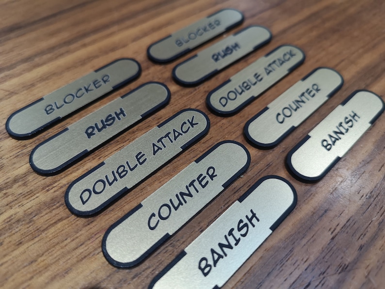 K&ouml;nnte beinhalten: Goldfarbene, rechteckige Spielmarken mit schwarzen R&auml;ndern. Jede Marke tr&auml;gt ein anderes Wort in schwarzer Schrift: "BLOCKER", "RUSH", "DOUBLE ATTACK", "COUNTER" und "BANISH". Die Spielmarken liegen auf einer Holzoberfl&auml;che.