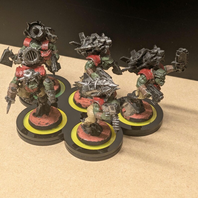 Peut inclure: Cinq figurines miniatures d'orcs &agrave; la peau verte, v&ecirc;tus d'une armure rouge et arm&eacute;s d'armes m&eacute;talliques. Ils sont plac&eacute;s sur des socles circulaires avec des accents jaunes et rouges, dispos&eacute;s sur une plateforme noire multi-socles. Les orcs sont d&eacute;taill&eacute;s avec diverses armes et &eacute;quipements.