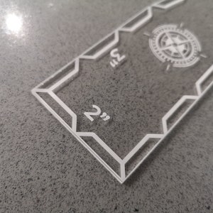 May include: Clear acrylic ruler with white markings and a compass rose design. The ruler measures up to 9 inches, with markings at 2 and 3 inches. The design features geometric shapes and a compass rose in the center.