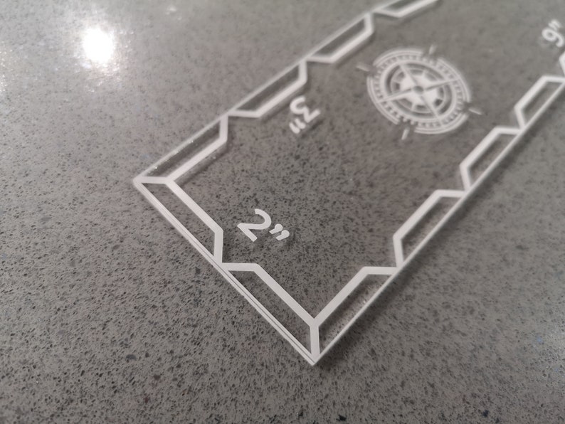 May include: Clear acrylic ruler with white markings and a compass rose design. The ruler measures up to 9 inches, with markings at 2 and 3 inches. The design features geometric shapes and a compass rose in the center.