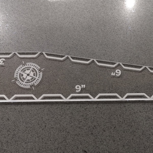 May include: Clear acrylic quilting ruler with a compass rose design. The ruler is 9 inches long and 2 inches wide at its widest point. It has measurements in inches and a decorative zigzag edge.