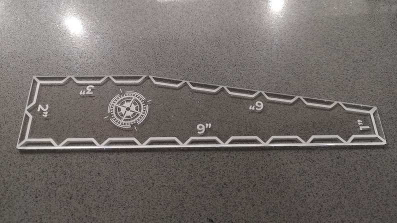 May include: Clear acrylic quilting ruler with a compass rose design. The ruler is 9 inches long and 2 inches wide at its widest point. It has measurements in inches and a decorative zigzag edge.