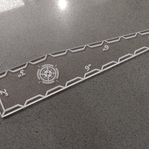 May include: Clear acrylic quilting ruler with etched markings and a compass rose design. The ruler is 9 inches long and features measurements from 2 to 7 inches. The edges have a decorative geometric pattern.