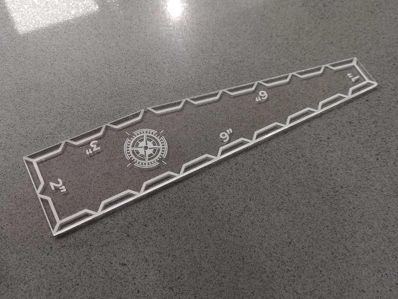 May include: Clear acrylic quilting ruler with etched markings and a compass rose design. The ruler is 9 inches long and features measurements from 2 to 7 inches. The edges have a decorative geometric pattern.