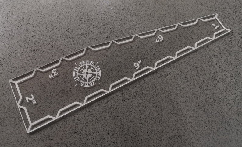 May include: Clear acrylic quilting ruler with a compass rose design and measurements in inches. The ruler is approximately 9 inches (22.86 cm) long and features angled edges and etched markings for precise cutting and alignment.