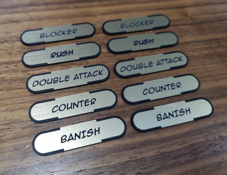 K&ouml;nnte beinhalten: Acht rechteckige, goldfarbene Spielmarken mit abgerundeten Kanten und schwarzen R&auml;ndern. Jede Marke hat ein anderes Wort in Schwarz aufgedruckt, darunter "BLOCKER", "RUSH", "DOUBLE ATTACK", "COUNTER" und "BANISH".