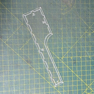 Puede incluir: Una plantilla de acrílico transparente para trabajos en cuero o costura, que presenta una sección superior rectangular con un recorte curvo y un mango largo y estrecho. La herramienta incluye marcas grabadas con los números 1, 2 y 3 visibles.