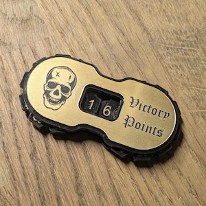 Puede incluir: Un contador de puntos de victoria de color latón con un gráfico de calavera y el número 16. Las palabras "Victory Points" están grabadas en el contador. El contador tiene un borde negro texturizado.