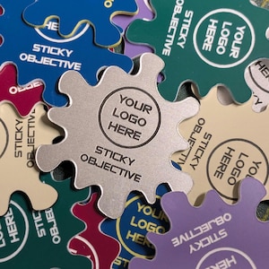 May include: A collection of colorful, gear-shaped stickers. The stickers are in various colors, including blue, green, purple, and beige. Each sticker has the text "STICKY OBJECTIVE" and "YOUR LOGO HERE" printed on it.