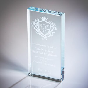 May include: A rectangular clear glass award with etched text and a decorative emblem. The text reads "Presented on behalf of The Northern Guild of Engineers" and "Stenning Academy". The award is approximately 6 inches tall.