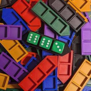 Op de afbeelding: Een verzameling veelkleurige 3D-geprinte plastic spelstukhouders in rood, blauw, groen, zwart, paars en oranje. Eén zwarte houder bevat drie groene zeszijdige dobbelstenen, elk met verschillende witte stippen op de bovenkant.