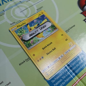 May include: A Pikachu Pok&eacute;mon trading card with a yellow background and the text "ABILITY USED" on a silver bar. The card features the character Pikachu and various game details.