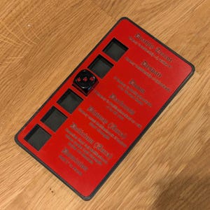 Op de afbeelding: Een rood en zwart tafelspelaccessoire met een dobbelsteen en tekst. De rode basis heeft uitsparingen en tekst, waaronder "Death Terror" en "Despair". Een zwarte dobbelsteen met drie stippen ligt in een uitsparing.