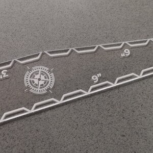 May include: Clear acrylic ruler with etched markings and a compass rose design. The ruler measures up to 9 inches, with markings at 1, 2, 3, 6, and 9 inches. The edges have a decorative geometric pattern.