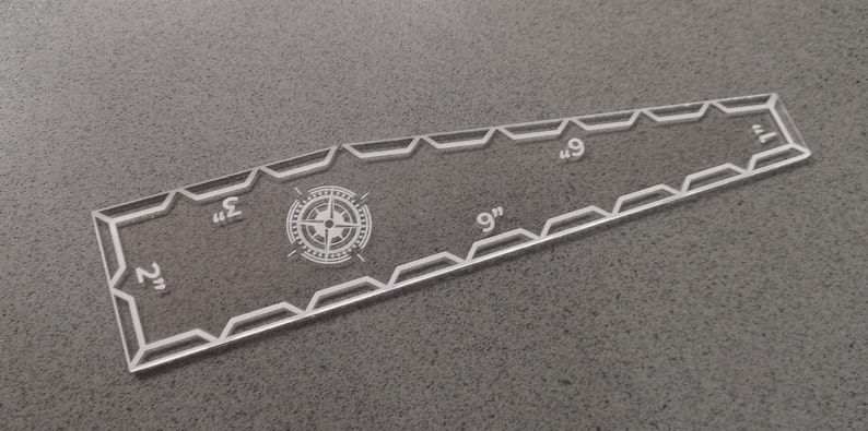 May include: Clear acrylic ruler with etched markings and a compass rose design. The ruler measures up to 9 inches, with markings at 1, 2, 3, 6, and 9 inches. The edges have a decorative geometric pattern.