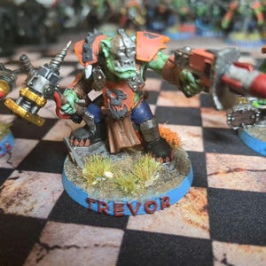 Op de afbeelding: Een miniatuur groene orkfiguur met oranje harnas, een blauwe broek en een bruine rok. De ork houdt een boor en een wapen vast. De basis is blauw met de naam "TREVOR" in rood. De figuur staat op een geruit oppervlak.