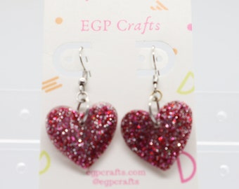 May include: A pair of pink glitter heart dangle earrings with silver hooks. The earrings are on a white card with the text "EGP Crafts" printed on it.