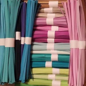 Puede incluir: Surtido de tiras de papel de colores para manualidades, dispuestas en paquetes y atadas con una cinta blanca. Las tiras son en tonos de verde, azul, rosa y marr&oacute;n.