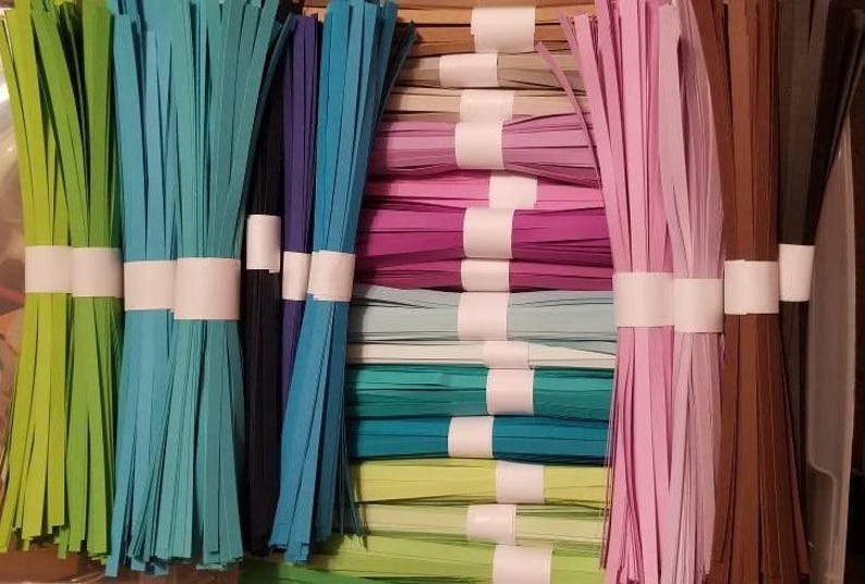 Puede incluir: Surtido de tiras de papel de colores para manualidades, dispuestas en paquetes y atadas con una cinta blanca. Las tiras son en tonos de verde, azul, rosa y marr&oacute;n.
