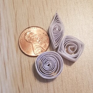 Puede incluir: Cuatro formas de papel blanco en espiral, incluyendo un diamante, un c&iacute;rculo y dos espirales, dispuestas sobre una superficie de madera. Tambi&eacute;n se puede ver una moneda de cobre en la imagen.