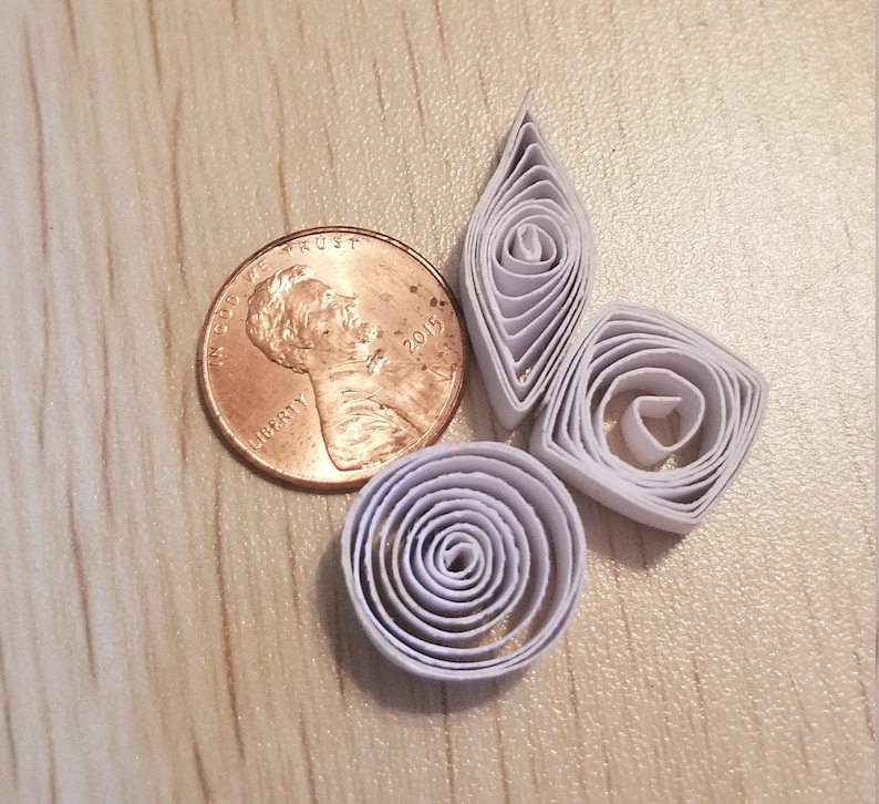 Puede incluir: Cuatro formas de papel blanco en espiral, incluyendo un diamante, un c&iacute;rculo y dos espirales, dispuestas sobre una superficie de madera. Tambi&eacute;n se puede ver una moneda de cobre en la imagen.
