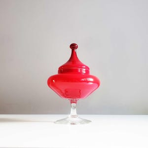Könnte beinhalten: Ein leuchtend rotes Glasgefäß mit Deckel, das auf einem Sockel aus klarem Glas steht. Das Gefäß hat einen bauchigen Körper und einen hohen, konischen Deckel mit einem kleinen, runden Knauf. Das Objekt steht auf einer weißen Oberfläche.