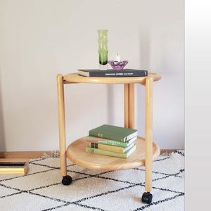 May include: A light wood, two-tiered side table with a round top and bottom shelf. The table has four legs and small black wheels. The top shelf holds a green glass vase, a purple glass dish, and a book. The bottom shelf holds a stack of books.
