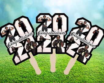 May include: Three black and white cardboard cutouts of the numbers 2021 with a football player in each number. The cutouts have wooden sticks attached to the back. The text "Congratulations" is written on each cutout.