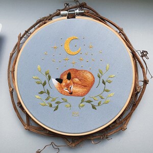 May include: Embroidered hoop art featuring a sleeping fox curled up under a crescent moon with stars. The fox is orange and brown, and the moon and stars are yellow. The hoop is light blue and has a wooden frame.
