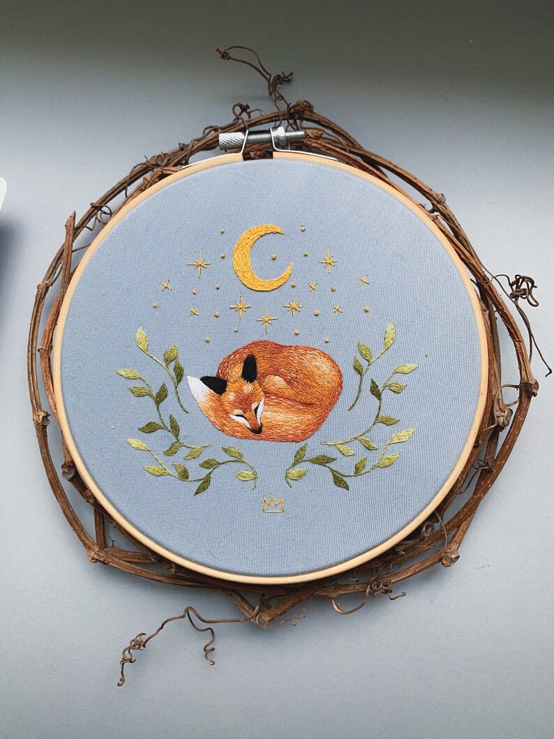 May include: Embroidered hoop art featuring a sleeping fox curled up under a crescent moon with stars. The fox is orange and brown, and the moon and stars are yellow. The hoop is light blue and has a wooden frame.