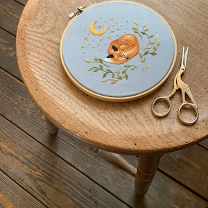 May include: A hand-stitched embroidery hoop with a sleeping fox, crescent moon, and stars on a light blue background. The hoop is on a wooden stool with gold embroidery scissors.