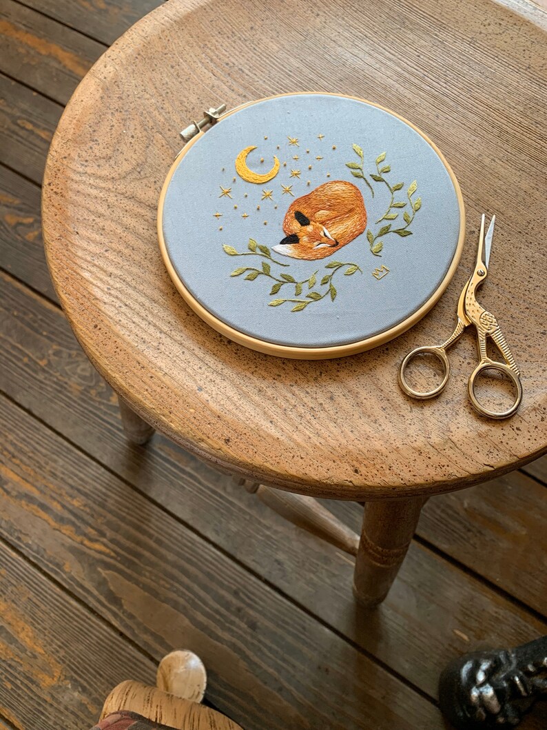 May include: A hand-stitched embroidery hoop with a sleeping fox, crescent moon, and stars on a light blue background. The hoop is on a wooden stool with gold embroidery scissors.