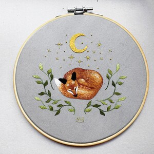 May include: Embroidered hoop art featuring a sleeping fox curled up under a crescent moon and stars. The fox is orange and brown, and the moon and stars are yellow. The hoop is surrounded by green leaves.