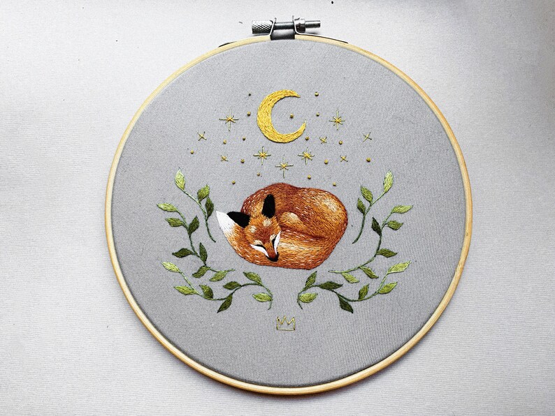 May include: Embroidered hoop art featuring a sleeping fox curled up under a crescent moon and stars. The fox is orange and brown, and the moon and stars are yellow. The hoop is surrounded by green leaves.