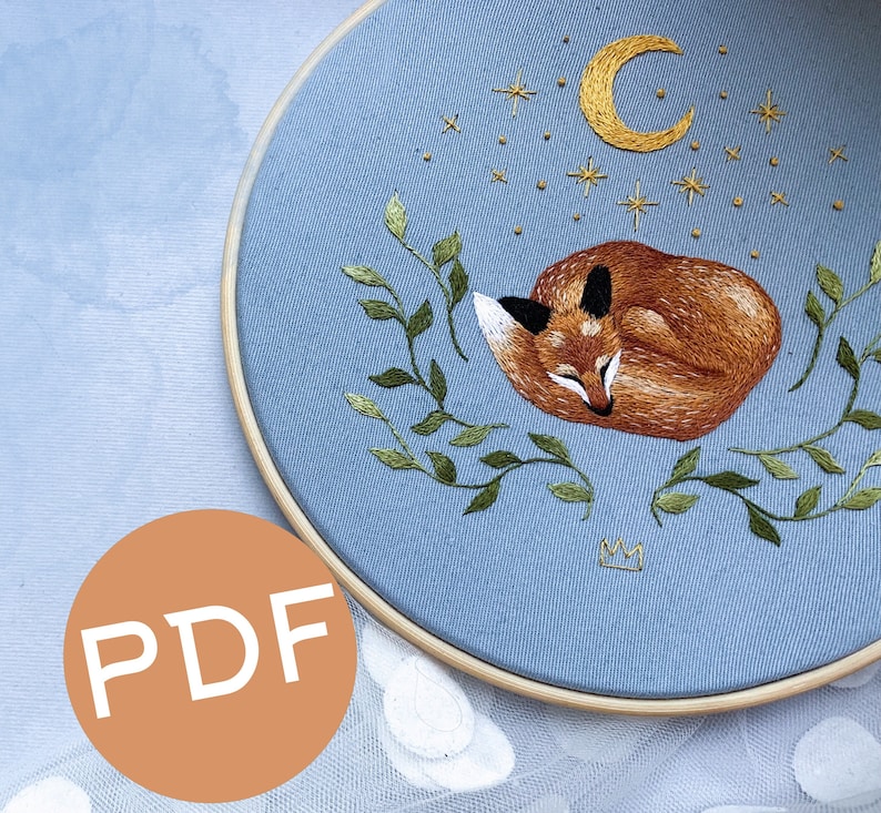 May include: Embroidered hoop art featuring a sleeping fox with a crescent moon and stars on a light blue background.