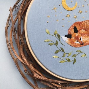 May include: Embroidered hoop art featuring a sleeping fox with a crescent moon and stars above. The fox is orange and brown, the moon is yellow, and the stars are gold. The background is light blue.