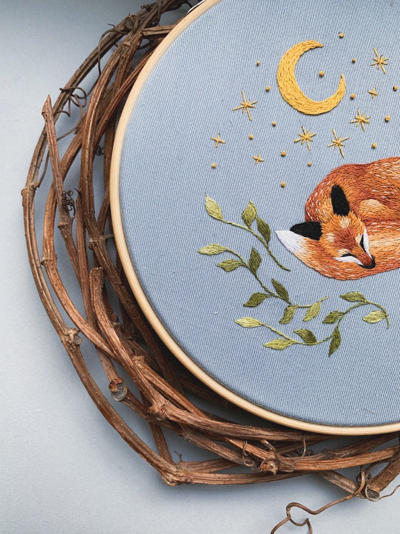 May include: Embroidered hoop art featuring a sleeping fox with a crescent moon and stars above. The fox is orange and brown, the moon is yellow, and the stars are gold. The background is light blue.