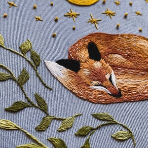 May include: Embroidered design of a sleeping fox with a crescent moon and stars above it. The fox is orange and brown with a white chest and black nose. The background is a light blue fabric with green leaves.