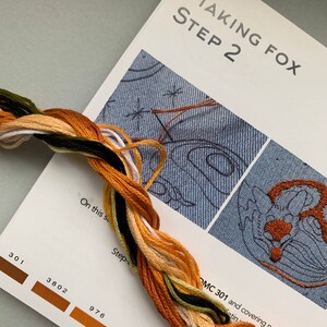 May include: A close-up of a page from a cross-stitch pattern book. The page shows a step-by-step guide for stitching a fox. The page includes a color chart with DMC floss numbers and a close-up of the stitching area. The text on the page reads "Making Fox Step 2" and "OMC 301 and covering needed zones (they're stitching using this pattern."