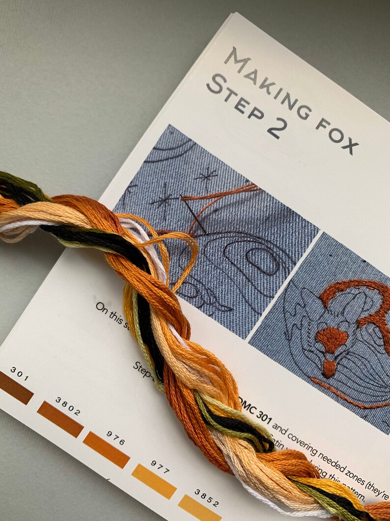 May include: A close-up of a page from a cross-stitch pattern book. The page shows a step-by-step guide for stitching a fox. The page includes a color chart with DMC floss numbers and a close-up of the stitching area. The text on the page reads "Making Fox Step 2" and "OMC 301 and covering needed zones (they're stitching using this pattern."