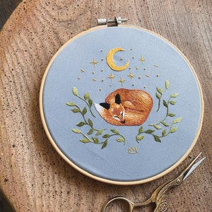 May include: Embroidered hoop art featuring a sleeping fox with a crescent moon and stars on a light blue background. The fox is surrounded by green leaves and a small crown is embroidered in the bottom center.