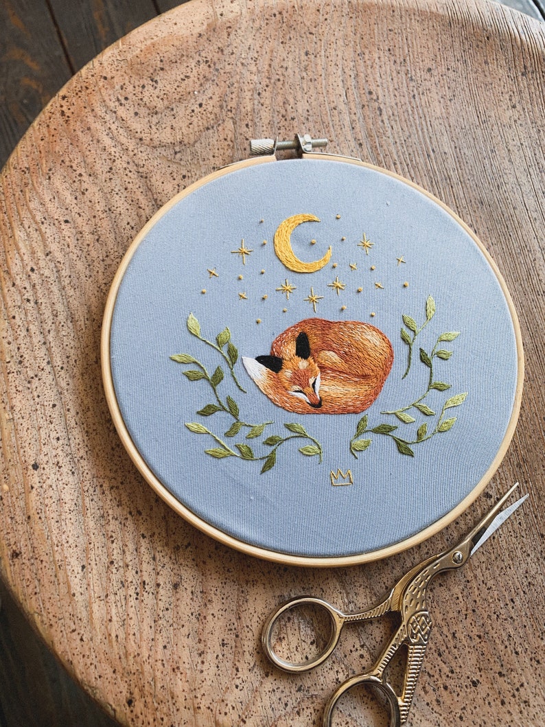 May include: Embroidered hoop art featuring a sleeping fox with a crescent moon and stars on a light blue background. The fox is surrounded by green leaves and a small crown is embroidered in the bottom center.