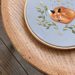 May include: A blue embroidery hoop with a sleeping fox, green leaves, a crescent moon, and gold stars on a light blue background.