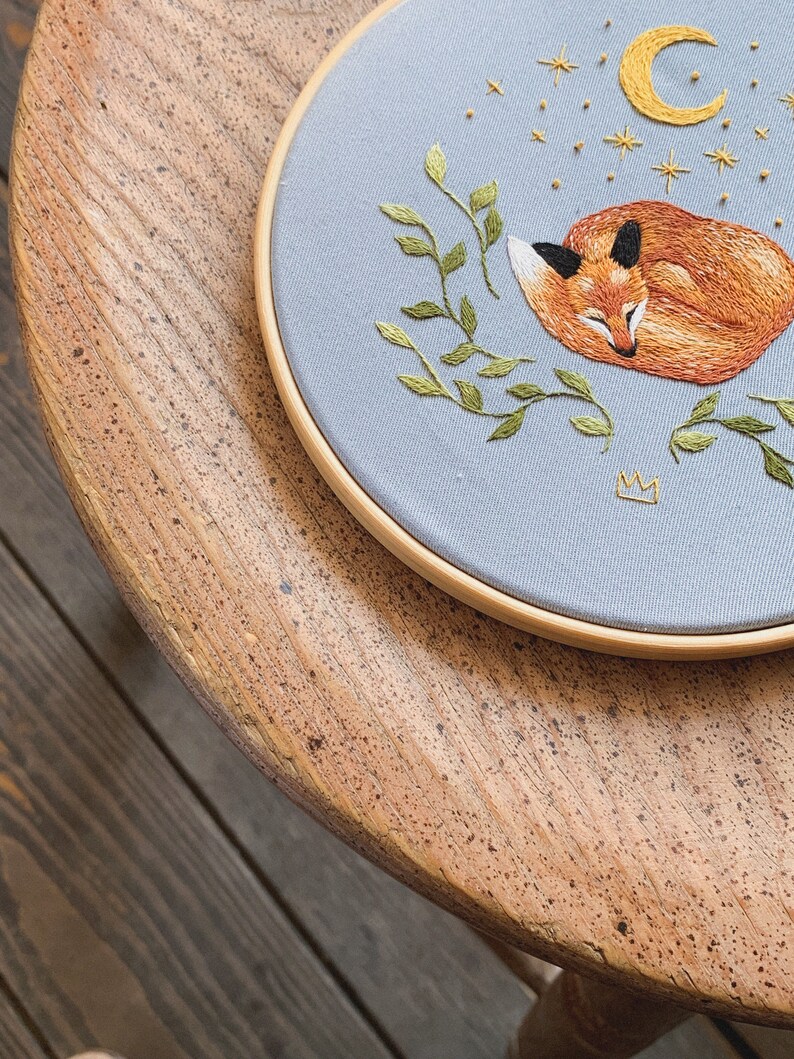May include: A blue embroidery hoop with a sleeping fox, green leaves, a crescent moon, and gold stars on a light blue background.