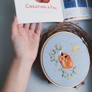 May include: A close-up of a completed embroidery hoop with a sleeping fox, crescent moon, and stars on a light blue fabric. The hoop is surrounded by a woven vine wreath. The text "Creating a Fox" is visible on a book in the background.