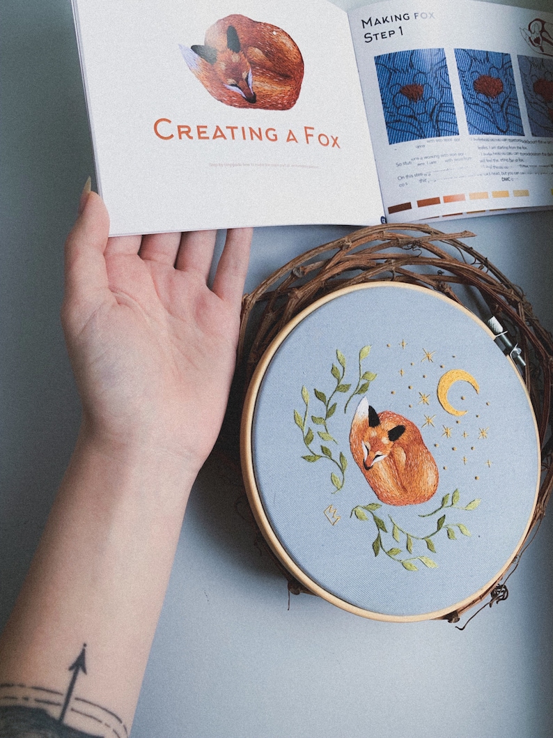 May include: A close-up of a completed embroidery hoop with a sleeping fox, crescent moon, and stars on a light blue fabric. The hoop is surrounded by a woven vine wreath. The text "Creating a Fox" is visible on a book in the background.