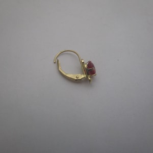 May include: A gold-colored earring with a red gemstone. The earring has a lever back closure. The gemstone is oval-shaped and set in a gold-colored metal. The earring is displayed against a white background.